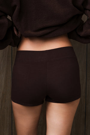 Rear view of a person wearing dark brown mini shorts and a matching dark brown top, standing against a dark wood plank background.
|M1