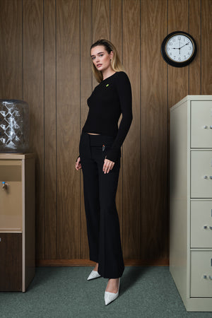 A woman stands in an office-like room with wood-paneled walls, wearing a fitted black long-sleeve top and black wide-leg pants with white pointed heels.
|M1