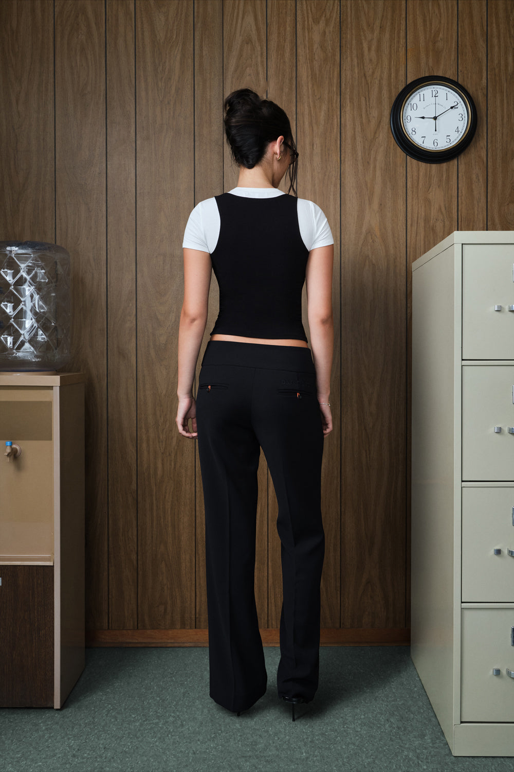 A woman stands with her back to the camera wearing a black tank over a white short-sleeve top and black wide-leg pants in an office setting with wood-paneled walls.
|M1