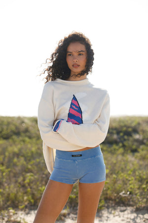 Woman wearing a white sweatshirt with a colorful design and blue shorts outdoors.
|M2
