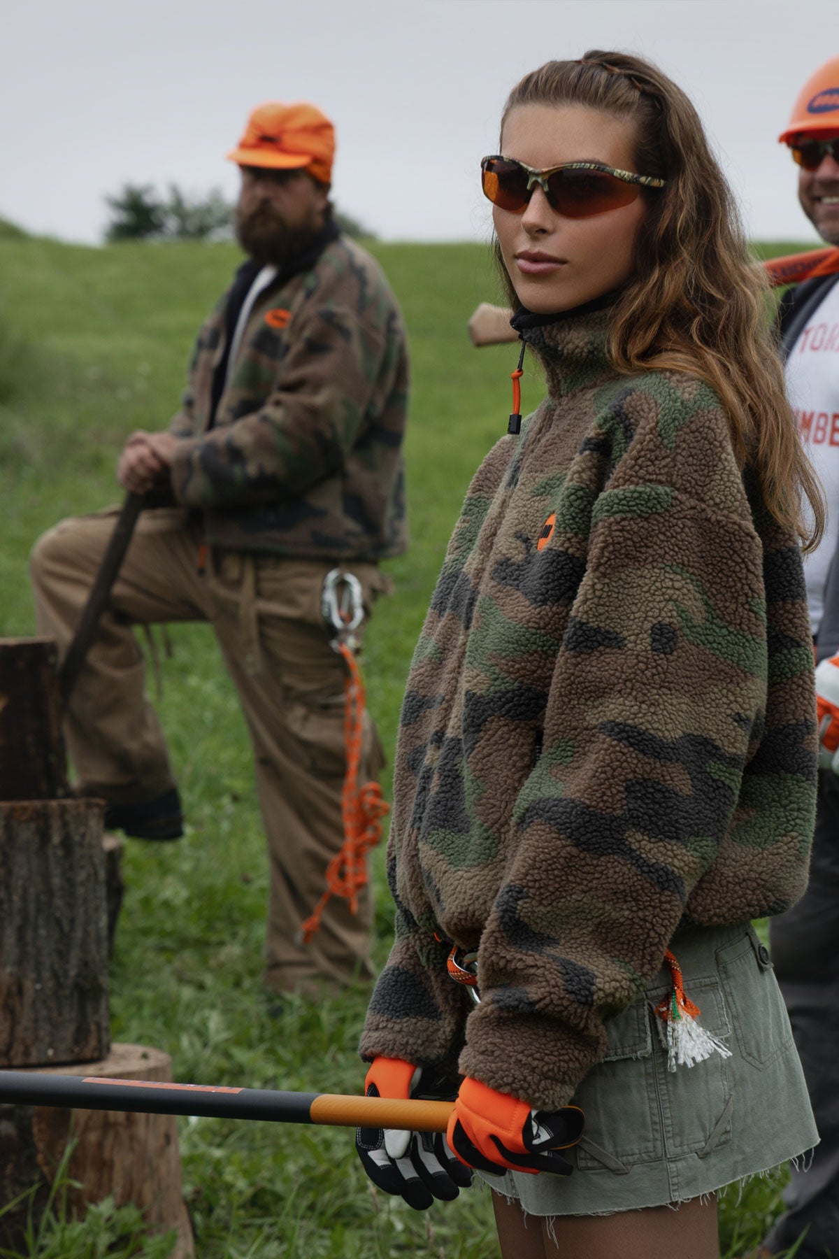 A woman in a woodland camouflage fleece pullover and sunglasses stands in a grassy outdoor setting holding a log splitter handle, with two men in camo clothing in the background.
|M2