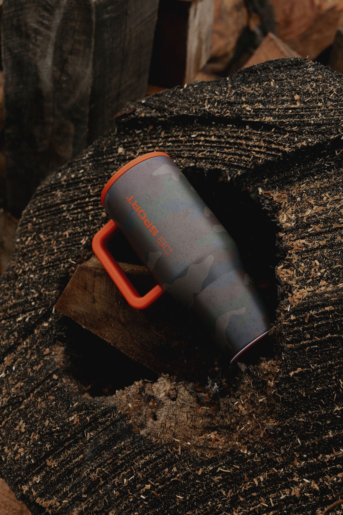 An insulated tumbler with a dark gray and black camouflage pattern and bright orange accents, resting on a textured dark wood surface.