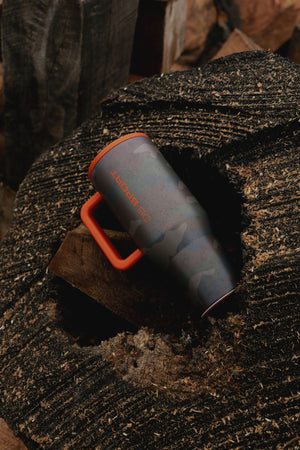 An insulated tumbler with a dark gray and black camouflage pattern and bright orange accents, resting on a textured dark wood surface.