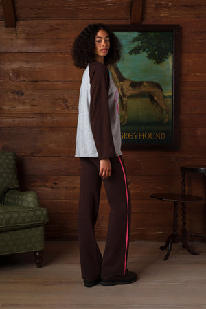 A woman stands in a rustic room with wood-paneled walls, wearing a raglan-style top with dark brown sleeves and light gray body panels, paired with dark brown pants featuring a bright pink stripe down the side. A framed painting of a greyhound is behind her.
|M1