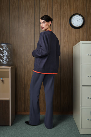 A woman stands in a retro office setting with wood-paneled walls, wearing a dark grey sweater with a bright orange hem trim and matching grey trousers.
|M1