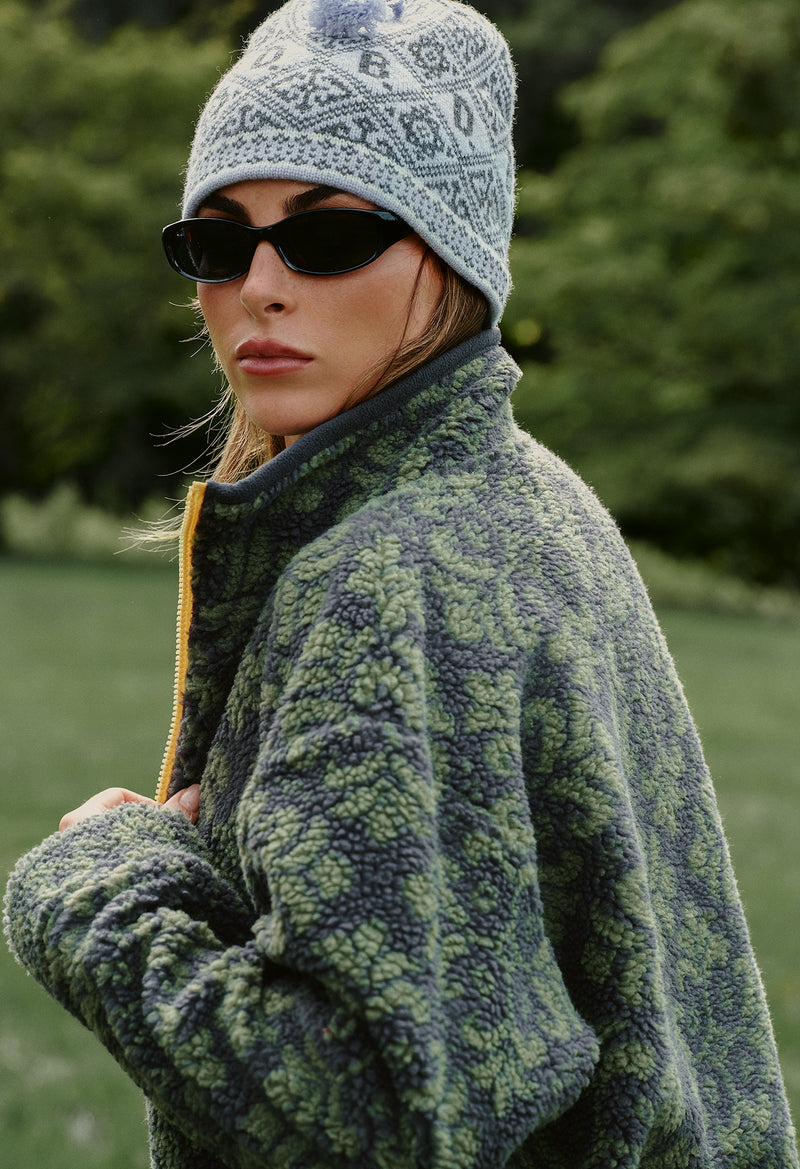 A woman wearing a green and gray patterned fleece jacket, a gray knit beanie, and black sunglasses, photographed outdoors against a blurred green background.