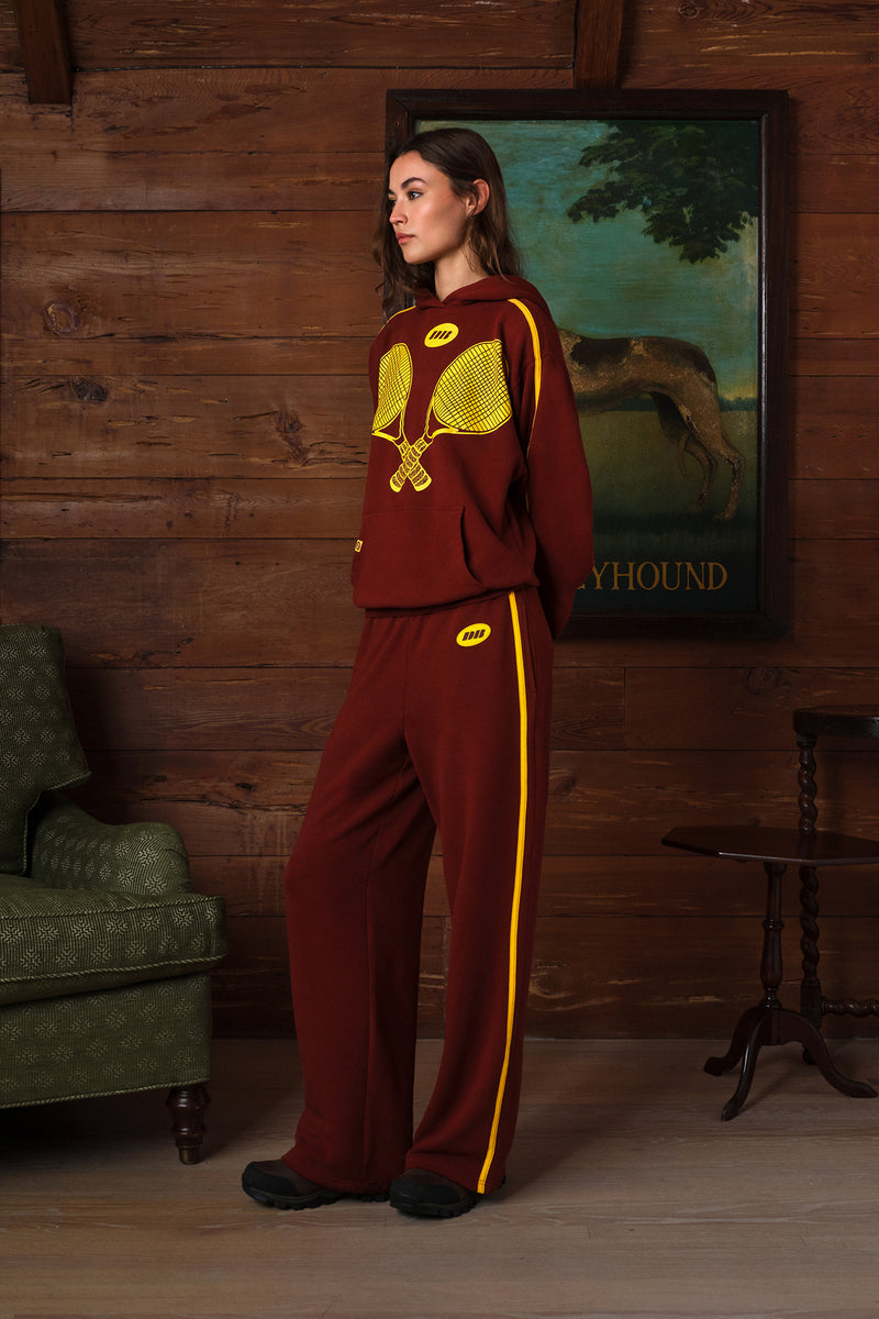 A woman stands in a rustic room with wood-paneled walls, wearing a burgundy sweatsuit with yellow accents. The set includes a cropped long-sleeve top with a yellow tennis racket graphic on the chest and matching wide-leg pants with a yellow side stripe.
|M1