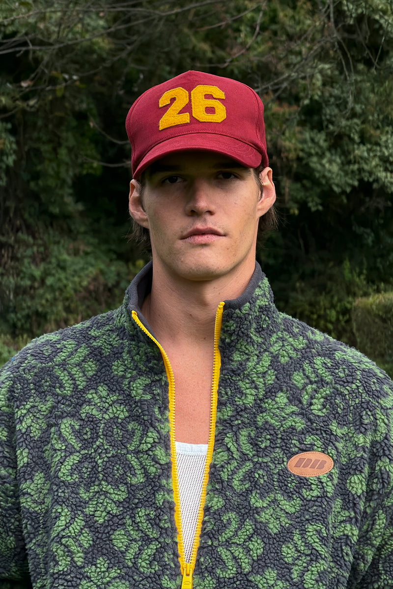 A man wearing a red baseball cap with yellow '26' and a green patterned fleece jacket with a yellow zipper, photographed outdoors against a blurred forest background.