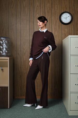 A woman models a matching dark brown tracksuit consisting of a long-sleeve crewneck sweater and wide-leg pants, styled with a white collared shirt and white pointed-toe heels in an office setting with wood-paneled walls.
|M1