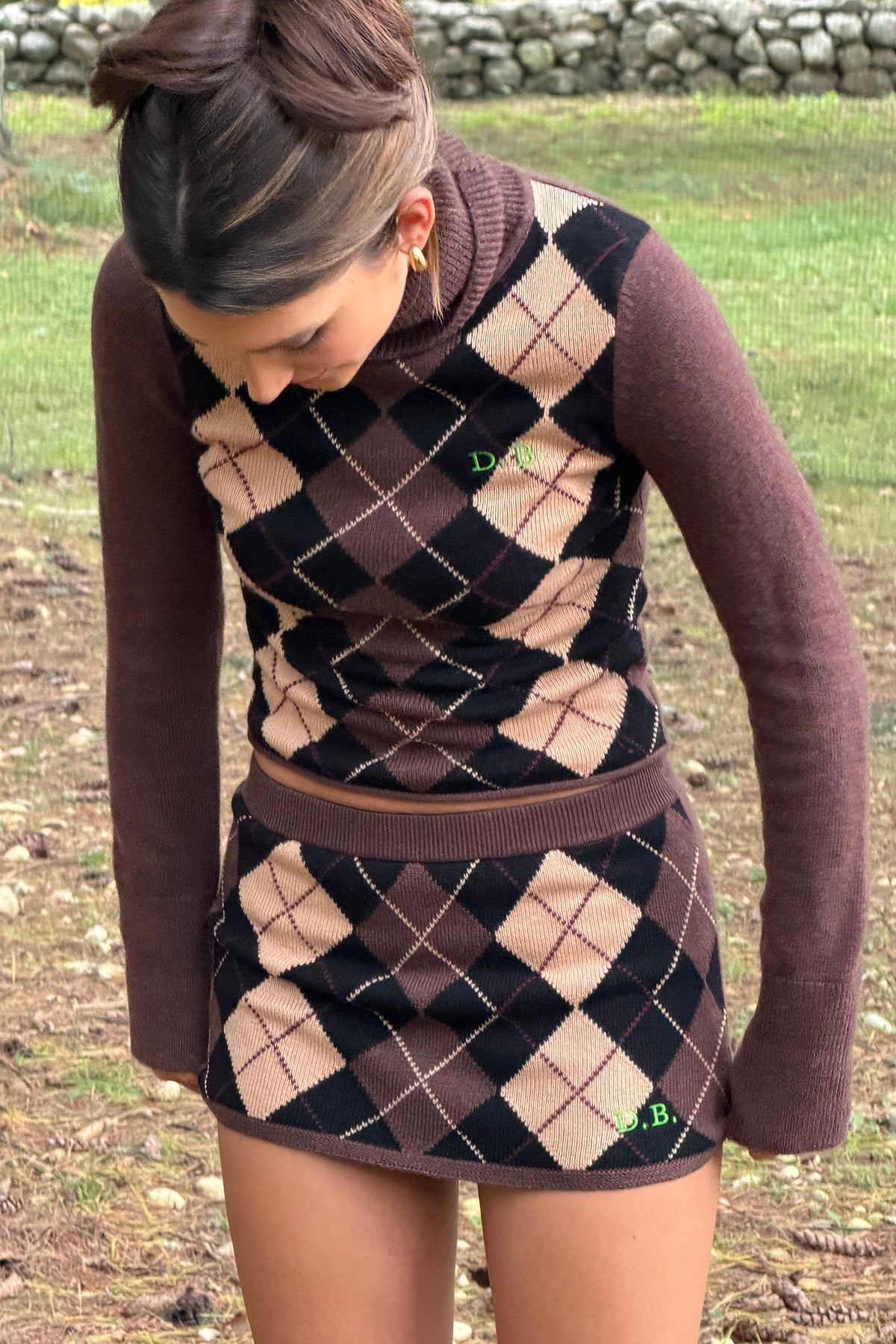 A woman wearing a brown and beige argyle knit turtleneck sweater vest and matching mini skirt, standing outdoors on a grassy ground.
|M2