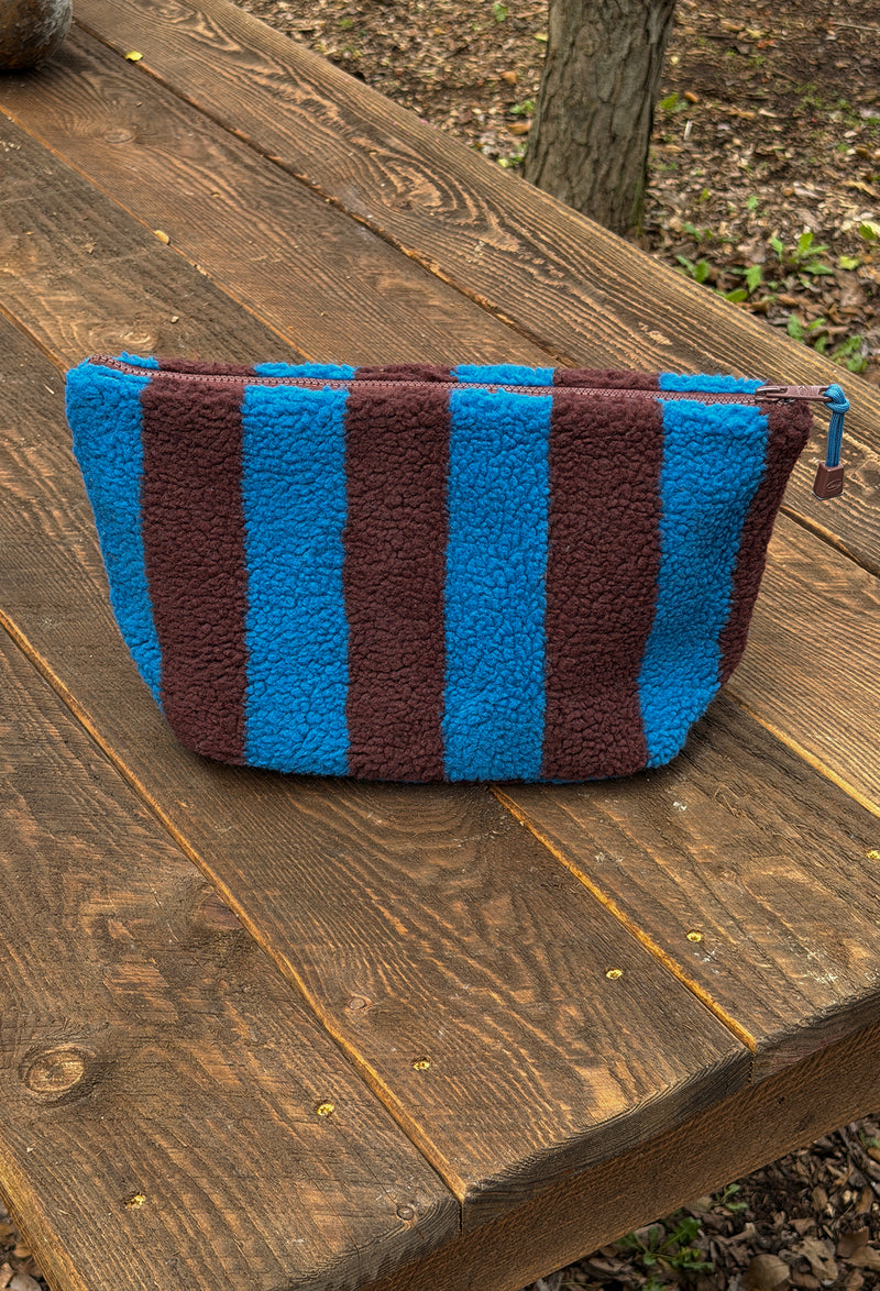 A zippered cosmetic pouch with vertical blue and dark brown stripes, made from a textured fleece-like fabric, resting on a rustic wooden table outdoors.