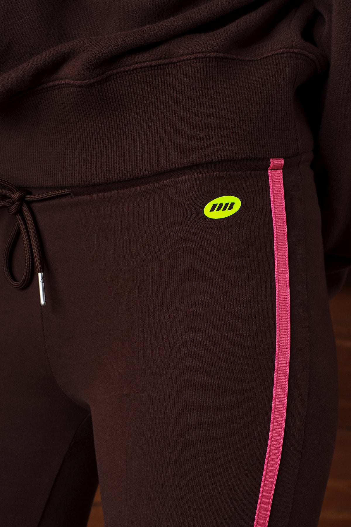 Close-up of dark brown sweatpants featuring a bright pink vertical stripe and a small lime green logo patch on the thigh.