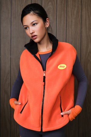 A woman wearing a bright orange fleece vest over a dark gray long-sleeve shirt, standing against a dark wood-paneled wall. The vest features a black zipper, black collar, and a small yellow oval logo on the left chest.
|M2