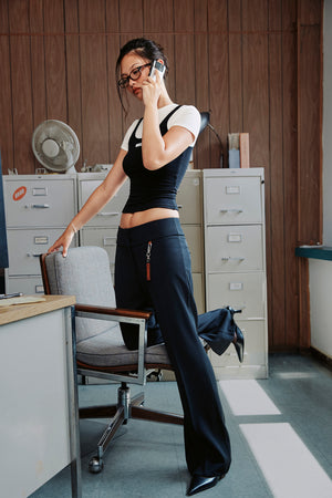 A woman in a black crop top and wide-leg navy pants stands in an office setting, leaning on a chair while talking on a phone.
|M1