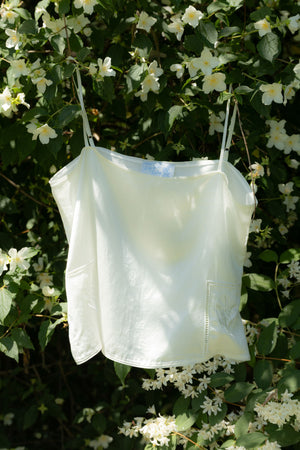 A white spaghetti-strap camisole top hanging outdoors against a background of green leaves and white flowers.