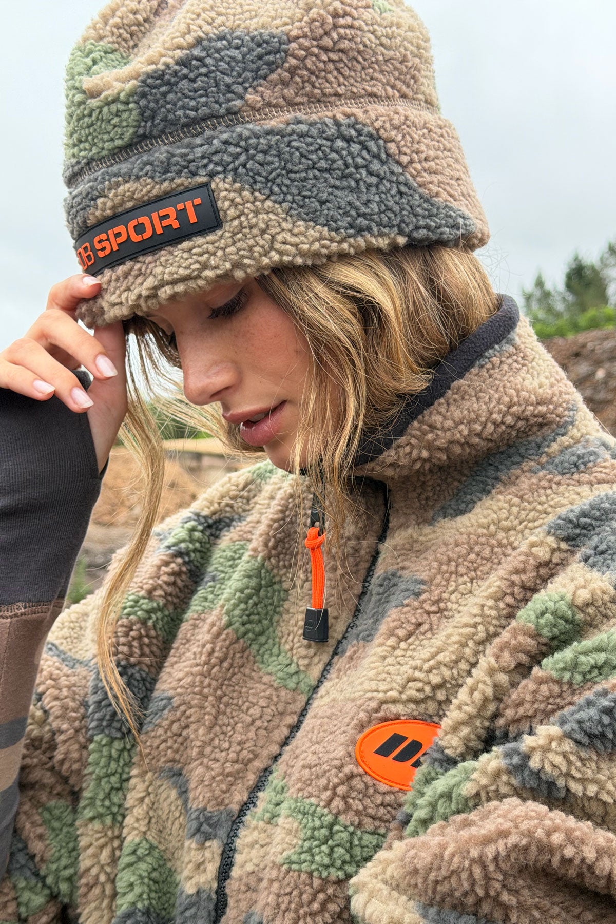 A woman wearing a beige, green, and brown camouflage fleece beanie with an orange and black 'DB SPORT' logo patch, paired with a matching camouflage fleece jacket with an orange logo patch on the chest.