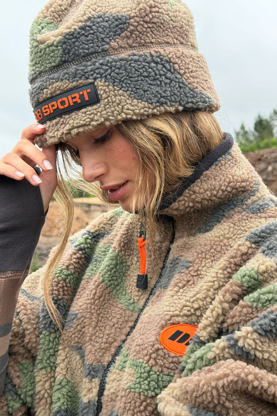 A woman wearing a beige, green, and brown camouflage fleece beanie with an orange and black 'DB SPORT' logo patch, paired with a matching camouflage fleece jacket with an orange logo patch on the chest.