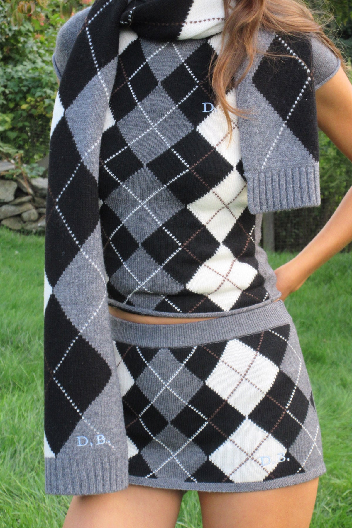 A person outdoors wearing a matching argyle knit short sleeve sweater, scarf, and mini skirt set in black, gray, and white, with small embroidered initials 'D.B.' visible on the scarf and shirt.
|M1