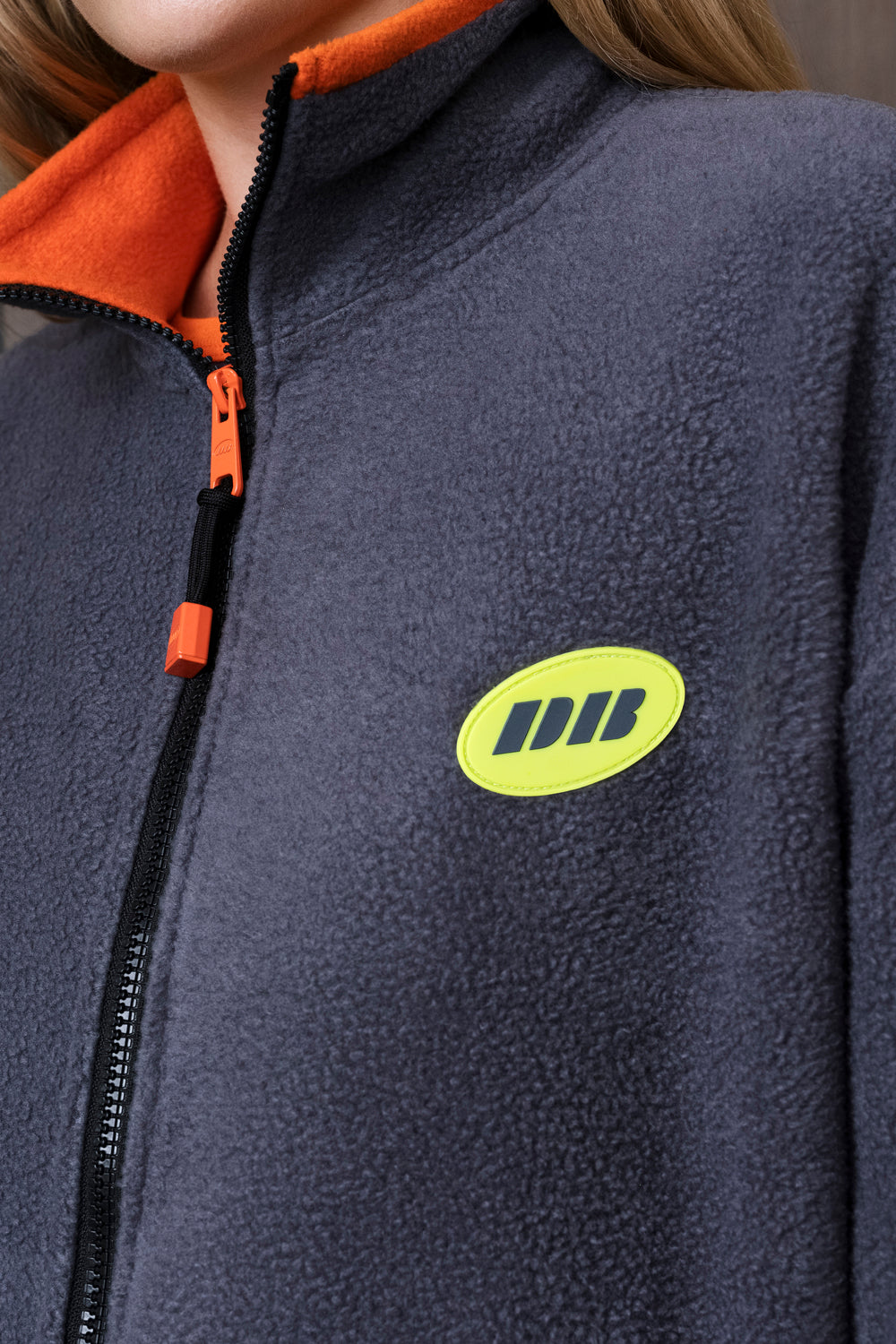 Close-up of a person wearing a dark gray fleece zip jacket with a bright orange collar and zipper pull, featuring a lime green oval logo patch on the chest.