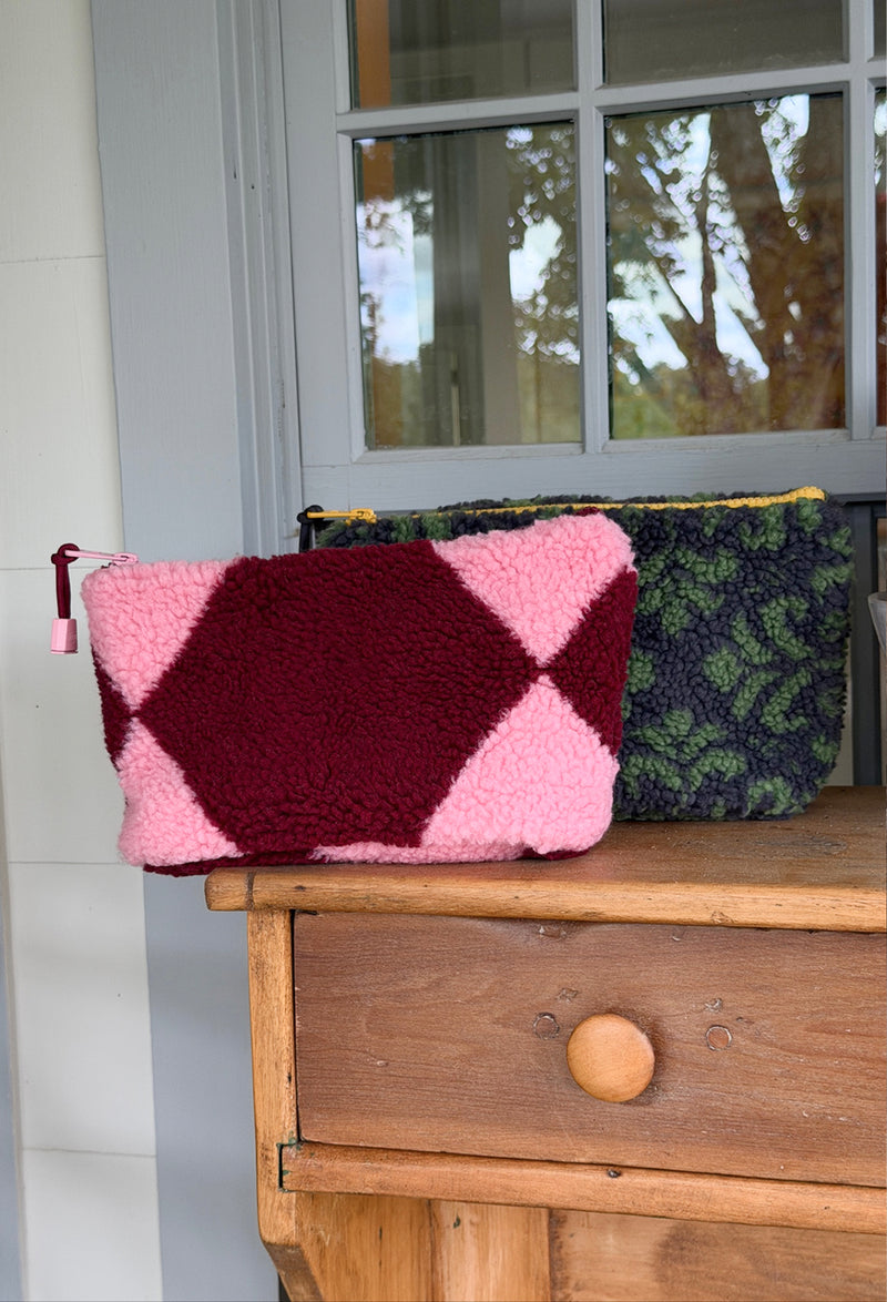 Two fleece fuzzy zippered pouches resting on a wooden chest in front of a window. The front pouch is made of pink and burgundy sherpa fabric, while the back pouch has a green and black leaf pattern.