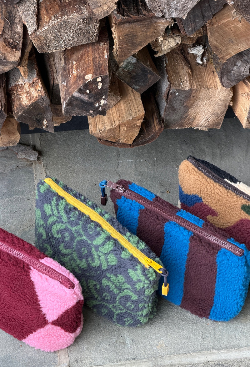 Three colorful fleece zippered pouches or small bags arranged on a stone surface in front of stacked firewood. The pouches have distinct patterns: one pink and burgundy geometric design, one green damask pattern on gray, and one blue and brown striped pattern.