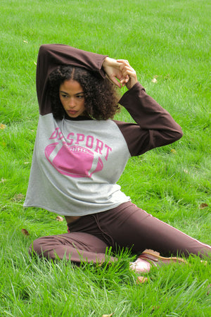 A woman sitting on a green grass lawn wearing a gray and brown raglan t-shirt with a pink football graphic and the word 'DB SPORT' on the front, paired with brown sweatpants.
|M1