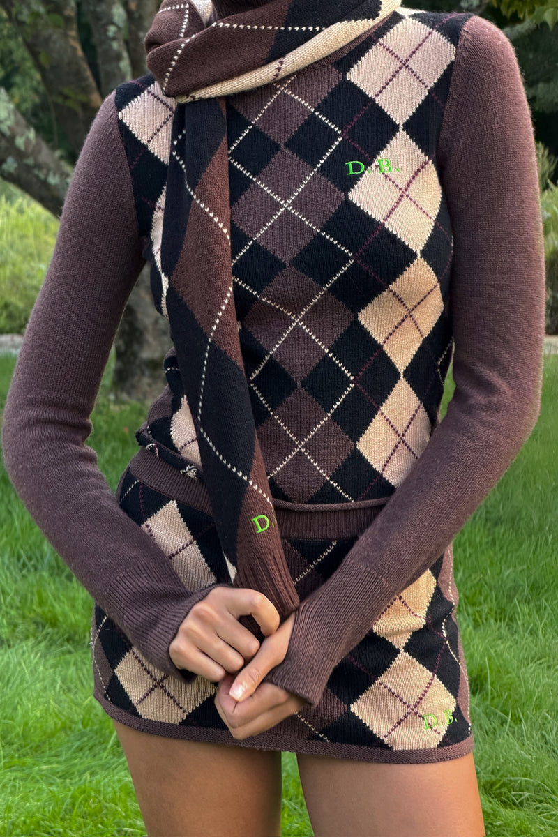 A person outdoors wearing a brown argyle sweater and matching knit scarf with grass visible in the background.