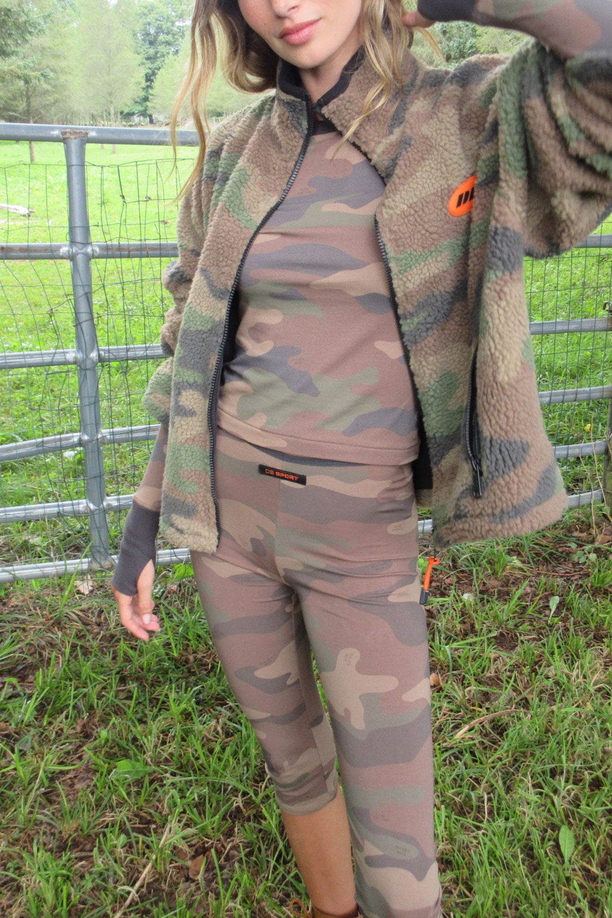 A woman wearing a matching camouflage fleece jacket, long-sleeve top, and leggings set, standing outdoors in a grassy field with a metal fence behind her.
|M1