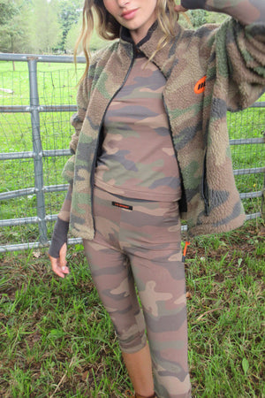 A woman wearing a matching camouflage fleece jacket, long-sleeve top, and leggings set, standing outdoors in a grassy field with a metal fence behind her.
|M1