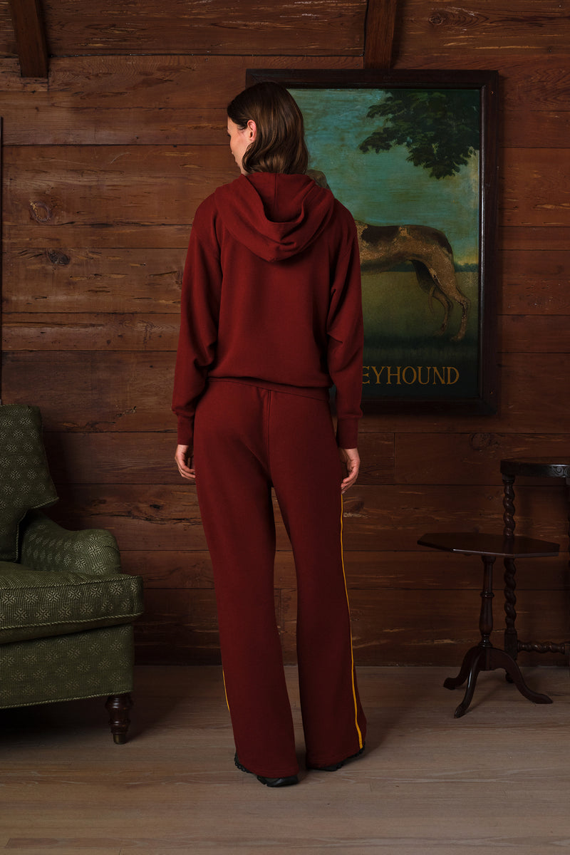 A woman stands with her back to the camera wearing a matching burgundy two-piece sweatsuit with a hooded sweatshirt and wide-leg pants. The room has rustic wood-paneled walls, a green armchair, a side table, and a framed painting of a greyhound.
|M1