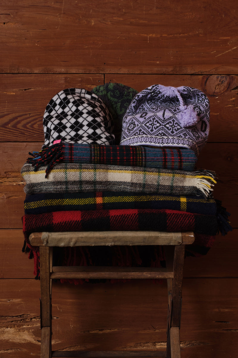 A stack of patterned knit hats and plaid scarves displayed on a rustic wooden stool against a dark wood plank wall.