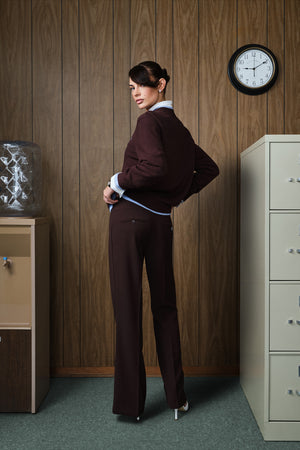 A woman stands in an office-like room with wood-paneled walls, a water dispenser, and a filing cabinet, wearing a dark brown tailored suit with a light blue shirt at the collar and cuffs.
|M1