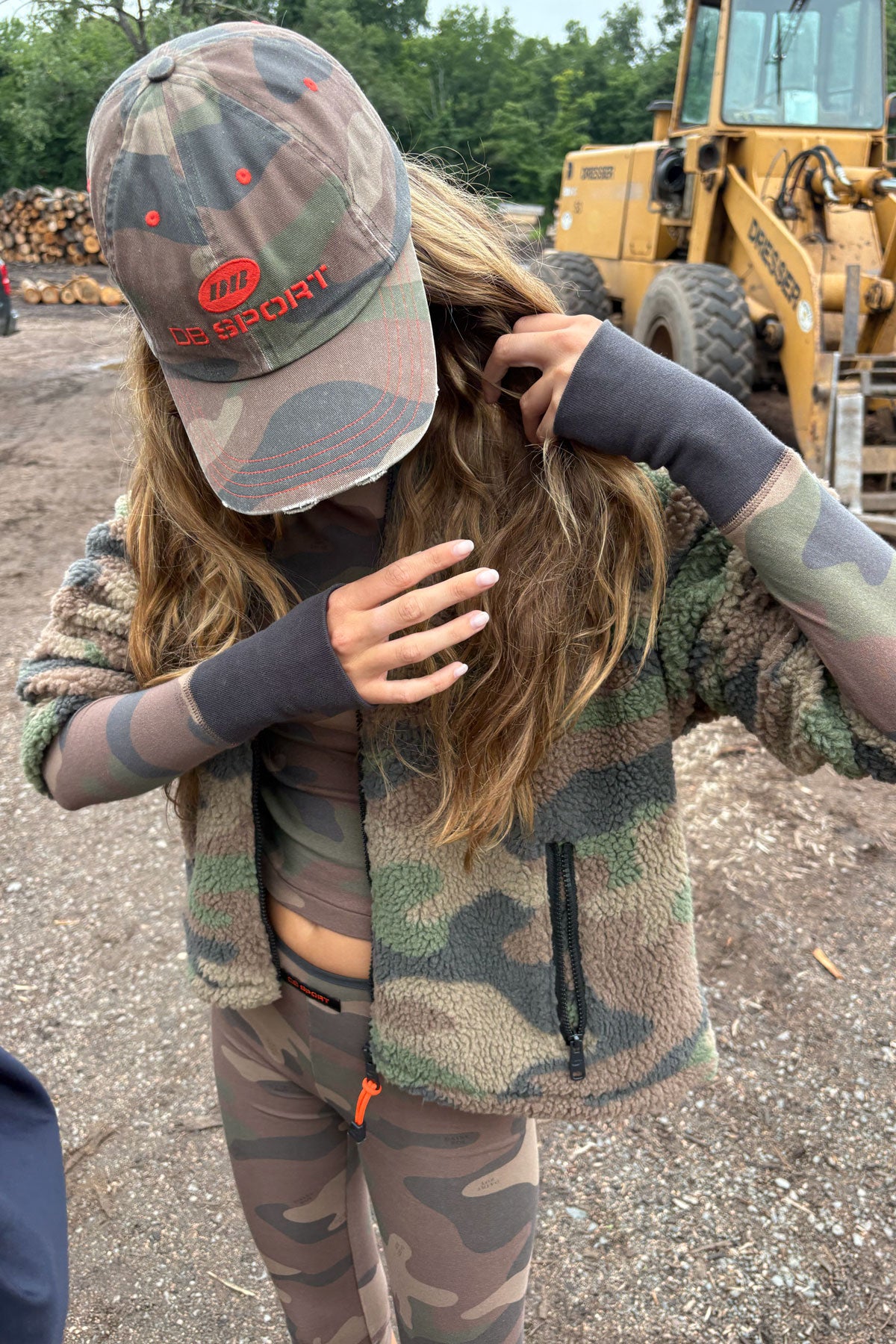 A person outdoors wearing a camouflage baseball cap with an orange 'DB SPORT' logo, a matching camouflage fleece jacket, and camouflage leggings. A yellow construction vehicle is visible in the background.