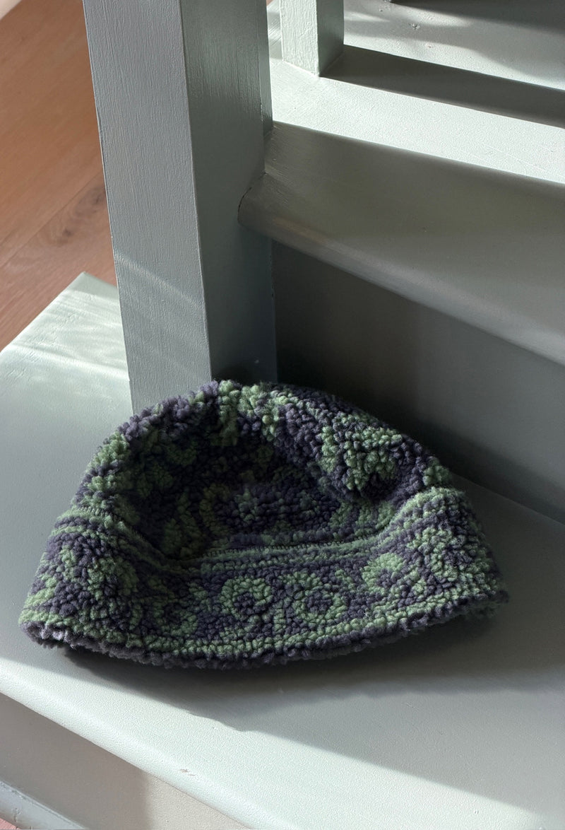 A textured, fuzzy knit beanie hat in a purple and sage green pattern, resting on a white wooden staircase step.