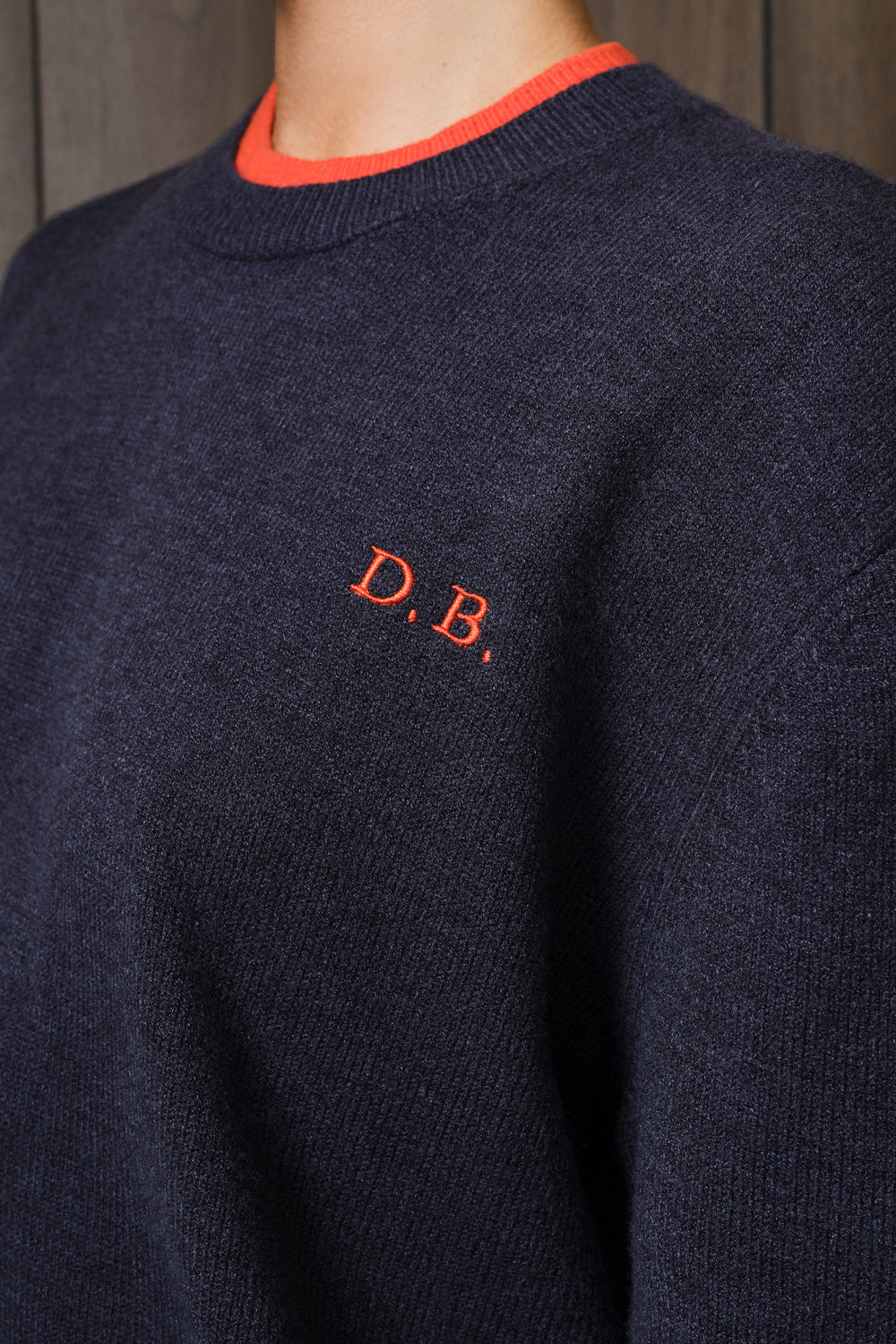Close-up of a navy knit sweater with a bright orange crew neck trim and an orange embroidered 'D.B.' logo on the chest.
|M1