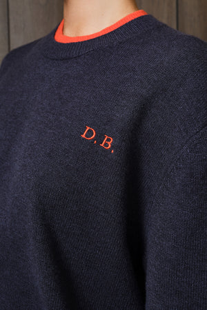 Close-up of a navy knit sweater with a bright orange crew neck trim and an orange embroidered 'D.B.' logo on the chest.
|M1