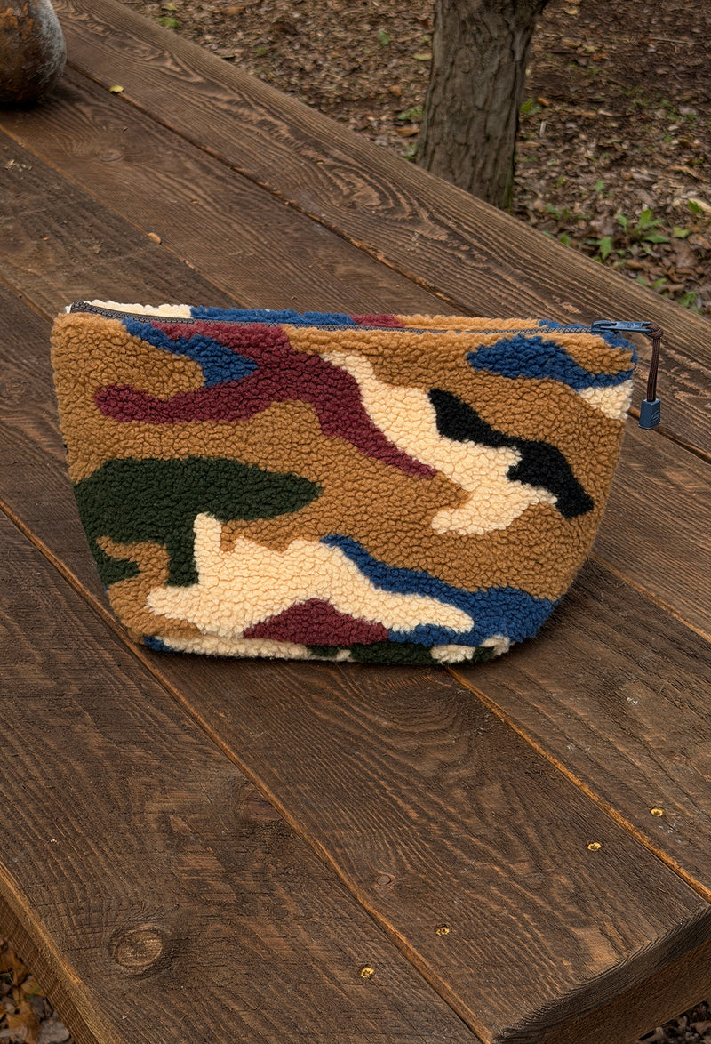 A fleece zip pouch with a multicolor camouflage pattern, resting on a rustic wooden deck outdoors.