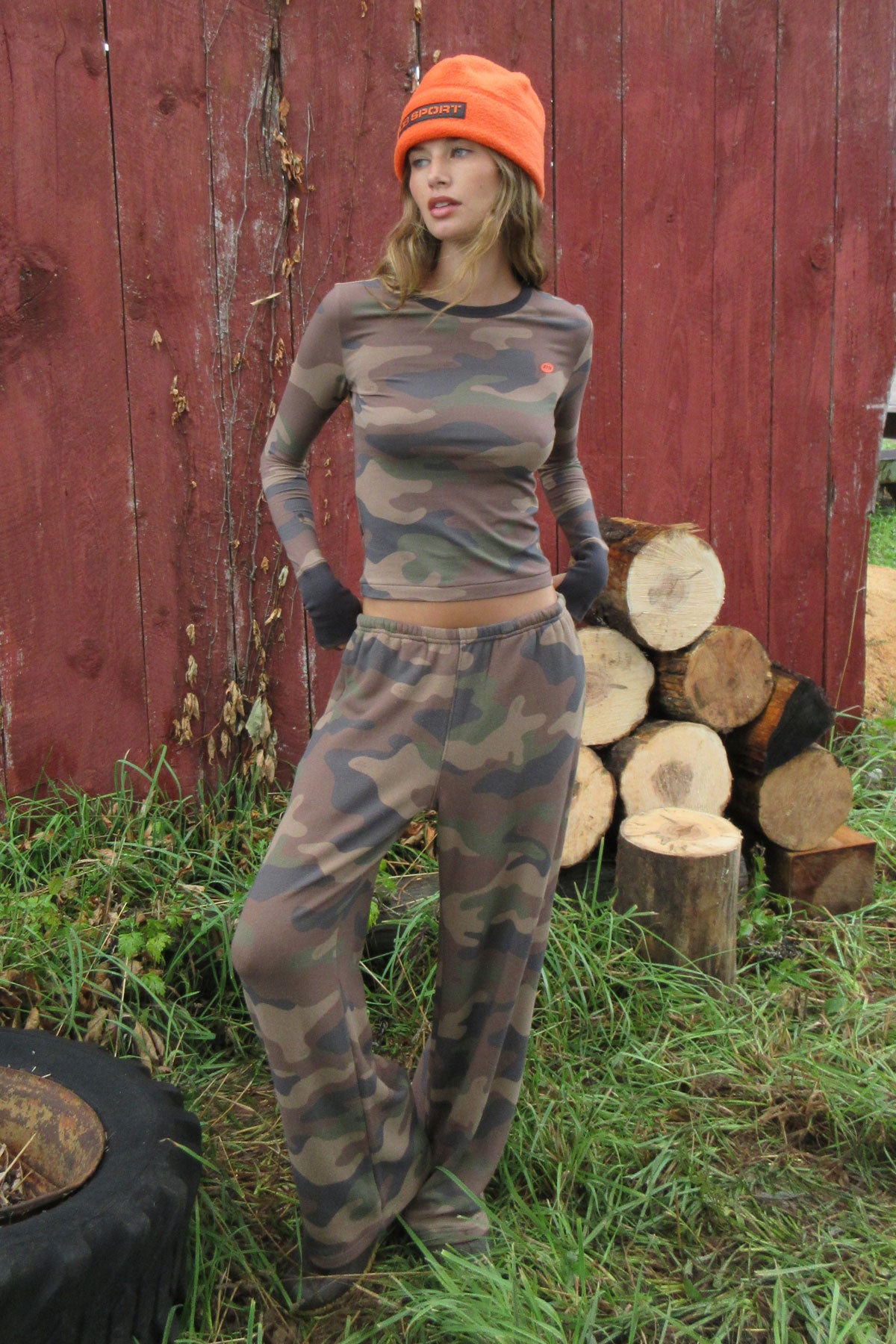 A woman wearing a camouflage long-sleeve crop top and matching camouflage pants, paired with an orange beanie and black gloves, standing outdoors in front of a red wooden fence with stacked logs.
|M1