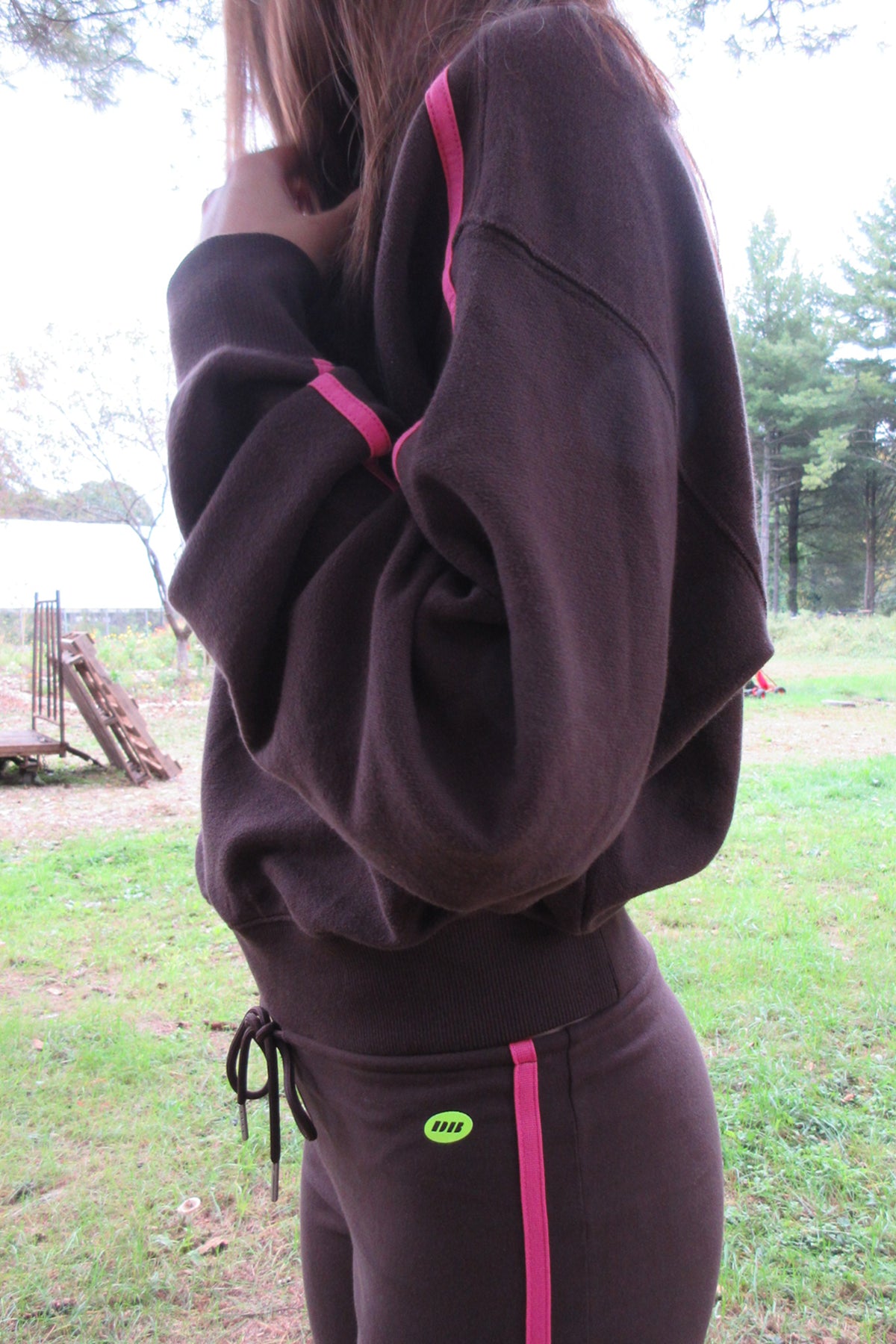A person outdoors wearing a dark brown sweatshirt and matching sweatpants with bright pink side stripes. The photo is cropped from the shoulders down, showing the back and side of the outfit, with grass and trees in the background.
|M1