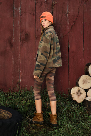 A woman stands outdoors in front of a red wooden fence and stacked logs, wearing a camouflage fleece jacket, matching camouflage capri leggings, an orange beanie, and brown boots.
|M1