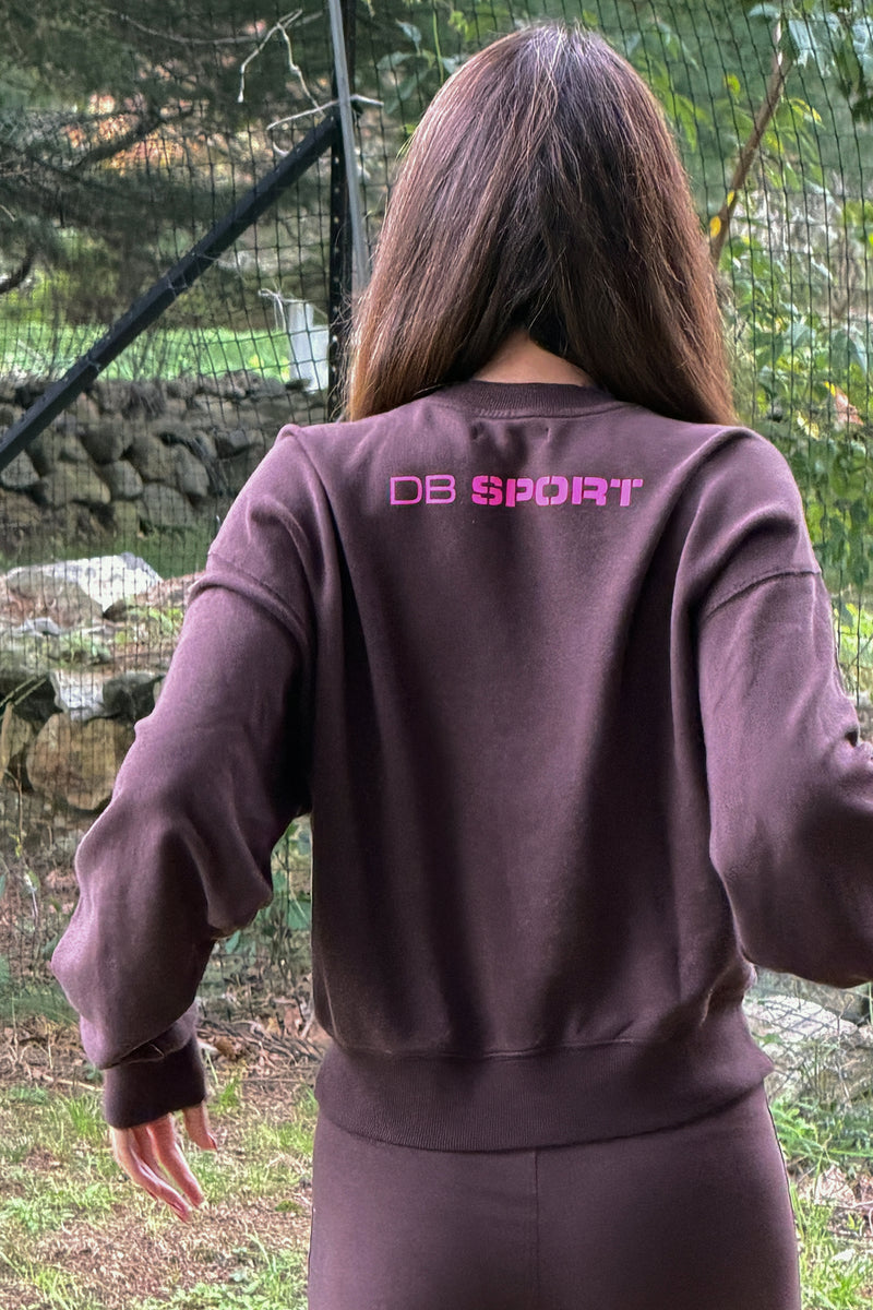 A person viewed from behind wearing a dark brown sweatshirt with 'DB SPORT' printed in pink across the upper back, set against an outdoor background with a stone wall and greenery.
|M1