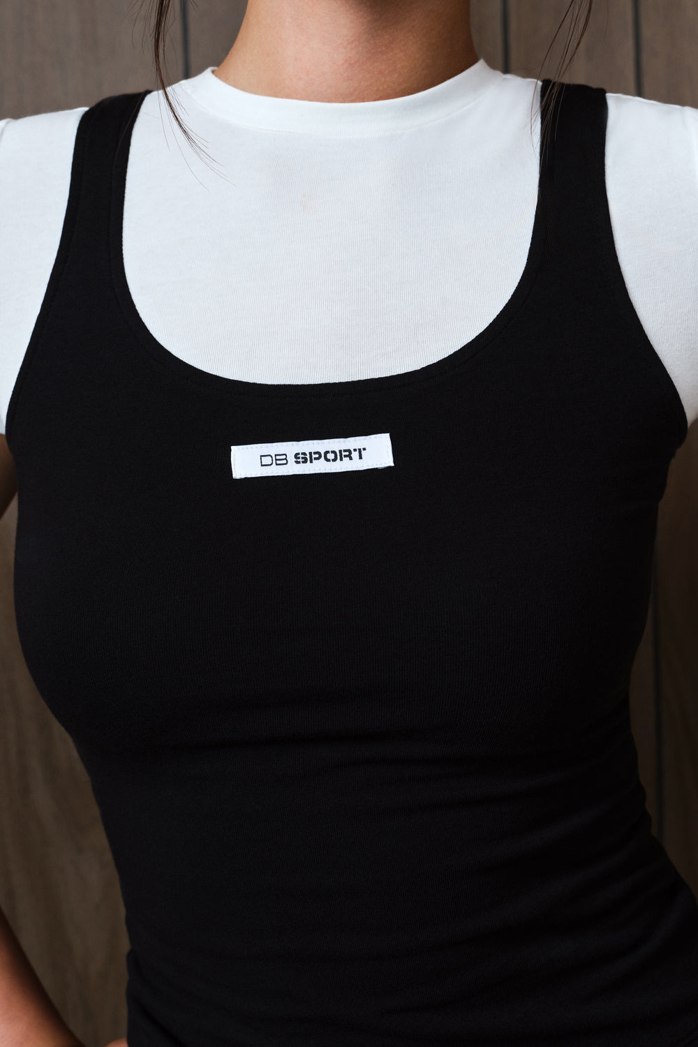 A close-up of a person wearing a black ribbed tank top over a white crewneck undershirt. The black tank top features a white rectangular chest label with the brand name 'DB SPORT'.