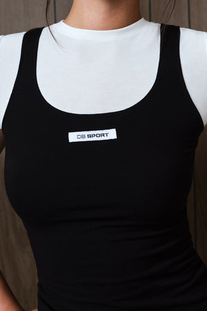 A close-up of a person wearing a black ribbed tank top over a white crewneck undershirt. The black tank top features a white rectangular chest label with the brand name 'DB SPORT'.