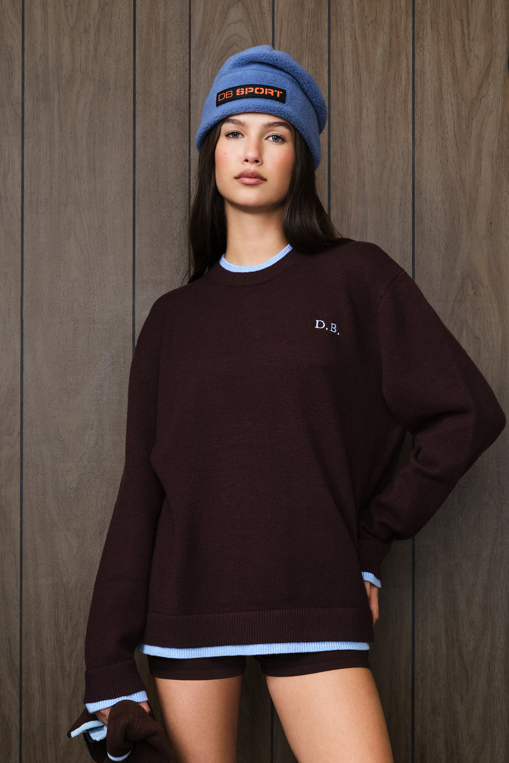 A woman wearing an oversized dark brown crewneck sweatshirt with light blue ribbed trim and a small white 'D.B.
|M2