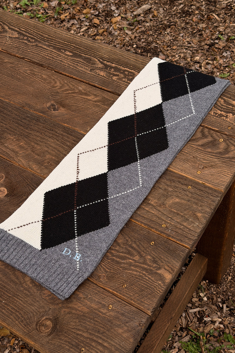 A knit scarf with an argyle pattern in black, off-white, and gray, laid out diagonally on a rustic wooden bench outdoors.