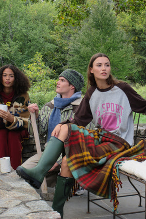Three models sitting outdoors on stone steps in front of a lush green forest. The woman on the right wears a gray and brown raglan sweatshirt with pink text, green rain boots, and a red, yellow, and green plaid blanket draped over her lap.
|M2