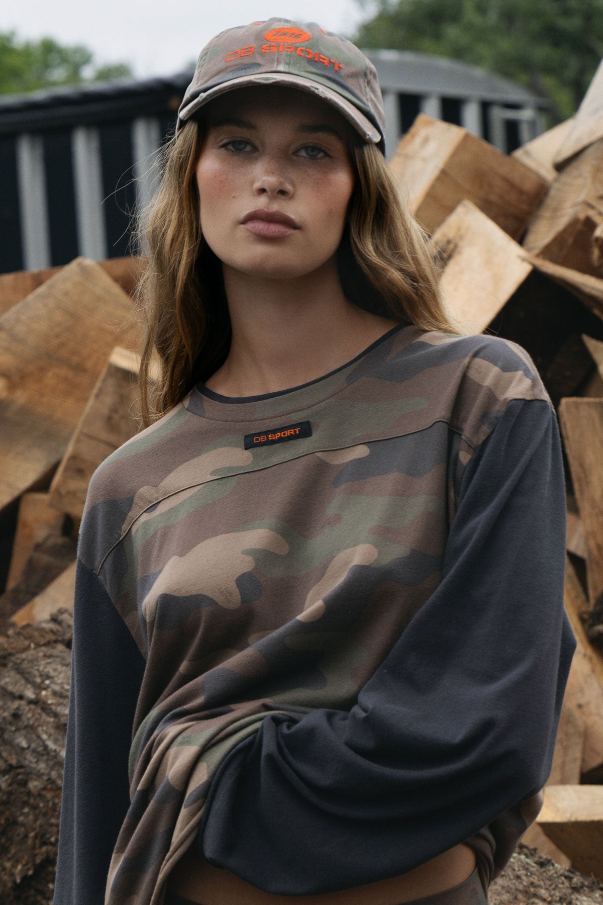 A woman wearing a camouflage long-sleeve crewneck shirt with dark grey sleeves and a matching camouflage baseball cap, standing in front of a pile of wood logs.
|M1