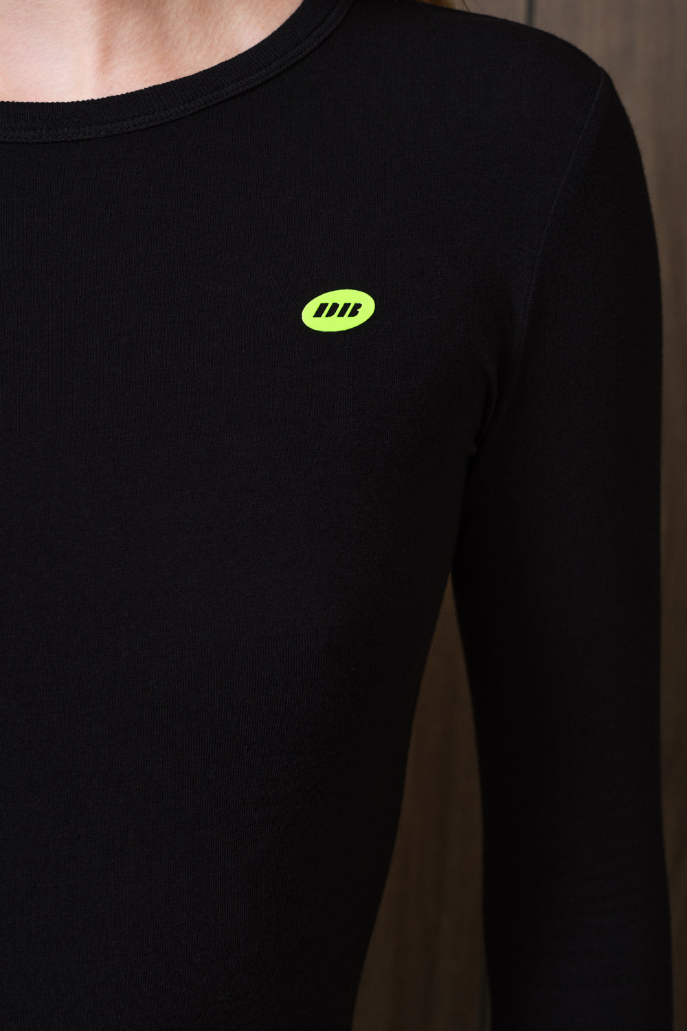 Close-up of a person wearing a black long-sleeve top with a small neon yellow-green oval chest logo featuring three diagonal bars.
|M1