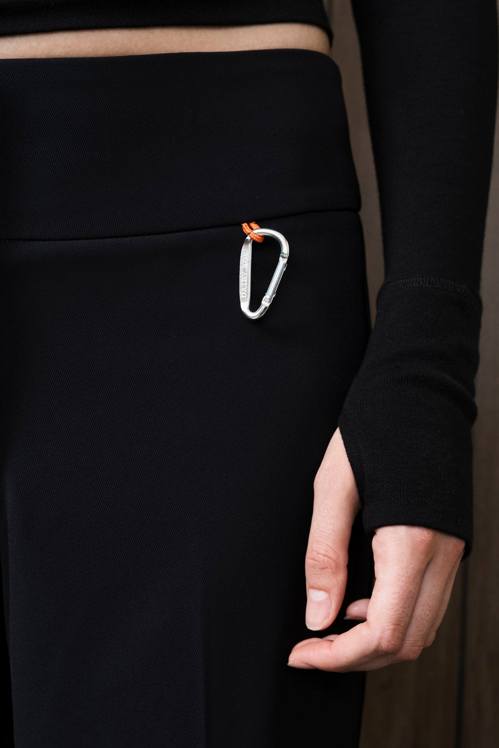 A close-up of a person wearing black pants and a black long-sleeve top, with a silver carabiner clipped to the side waistband. The carabiner has a small orange accent.
|M1