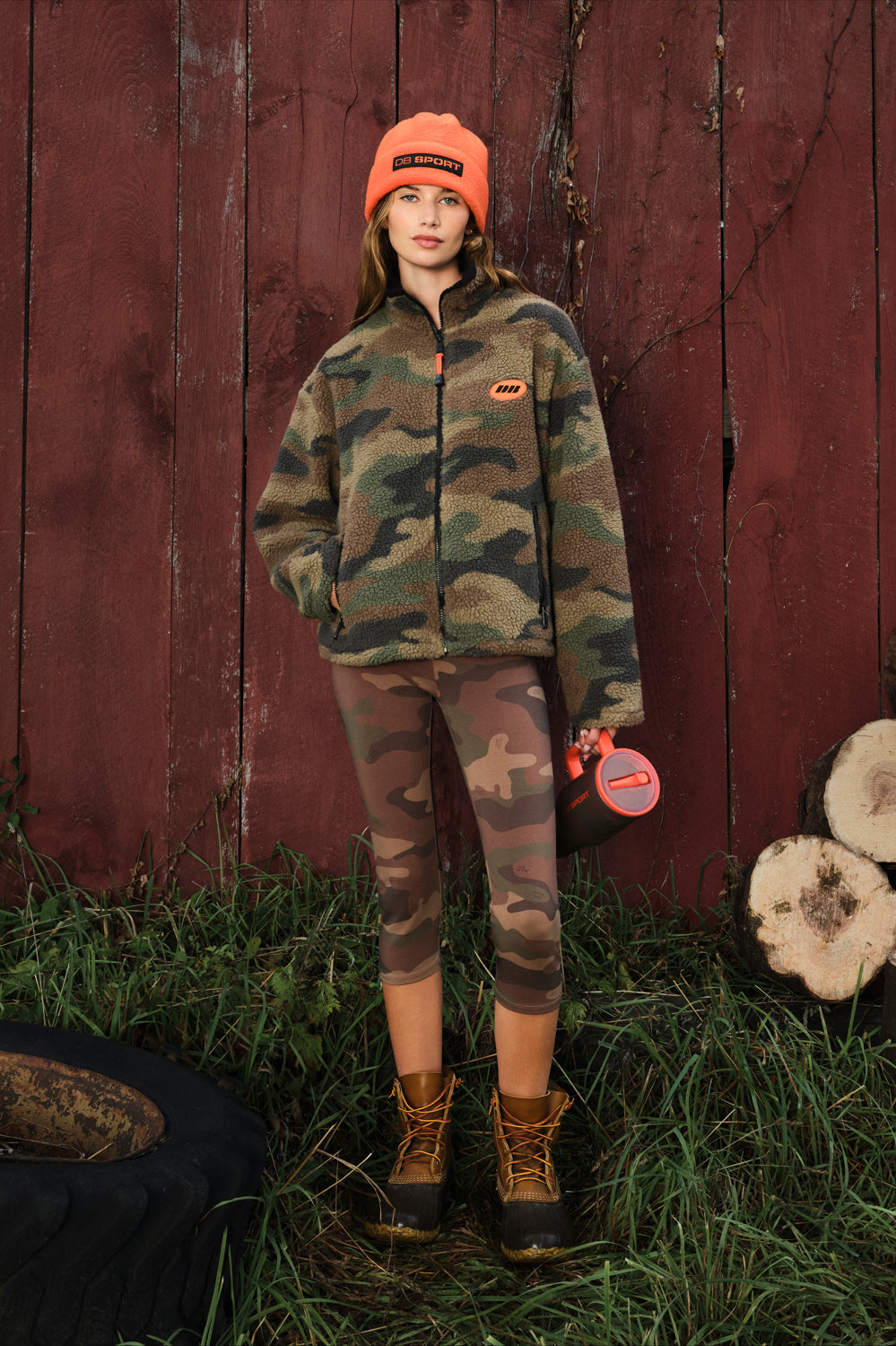 A model stands outdoors in front of a red wooden barn wall wearing a camouflage fleece jacket, matching camouflage leggings, brown hiking boots, and an orange beanie. A small orange logo patch is visible on the chest of the jacket.
|M1